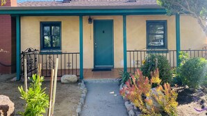 A single-story house with a green door, two windows, a small porch, and a garden with various plants. - 2 Bed 1 Bath  , Grand Pa's Cottage, Long Term Vacation Rentals . (Santa Barbara)