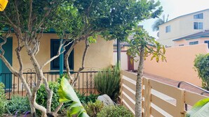 A residential area with a house featuring a beige exterior, a black metal fence, and a small garden with various plants and a tree. - 2 Bed 1 Bath  , Grand Pa's Cottage, Long Term Vacation Rentals . (Santa Barbara)