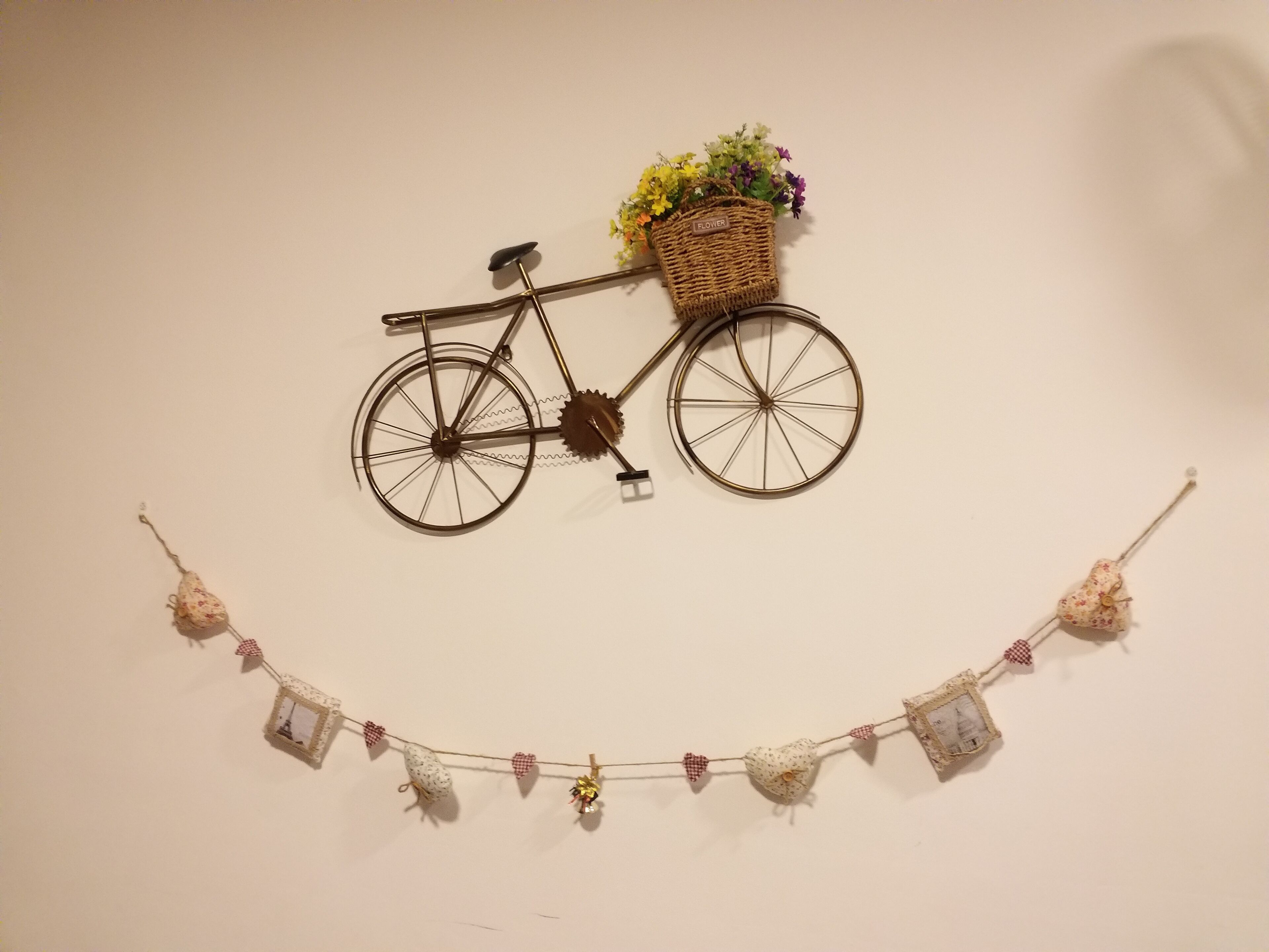 Une décoration murale représentant un vélo avec un panier rempli de fleurs et une guirlande d’ornements en forme de cœur.