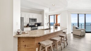 Une cuisine moderne dotée d’un grand îlot central, de tabourets de bar en bois et offrant une vue sur l’océan grâce à de grandes fenêtres.