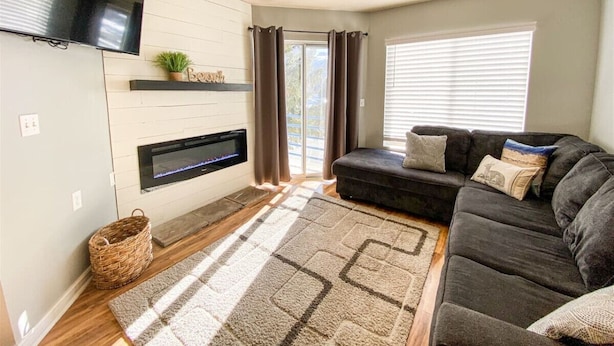 A modern living room with a flat-screen TV, fireplace, dark sofa, and large windows. - Beautiful Inn Building Condo #208 Pet friendly (Garden City)