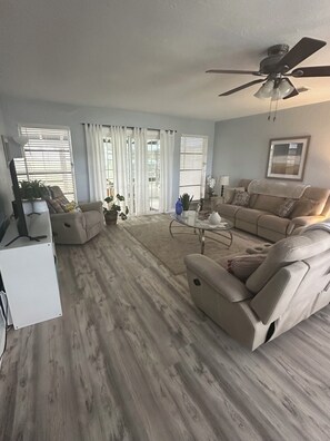 Ein geräumiges Wohnzimmer mit einem beigefarbenen Sofa, einem Glastisch und einem Deckenventilator.