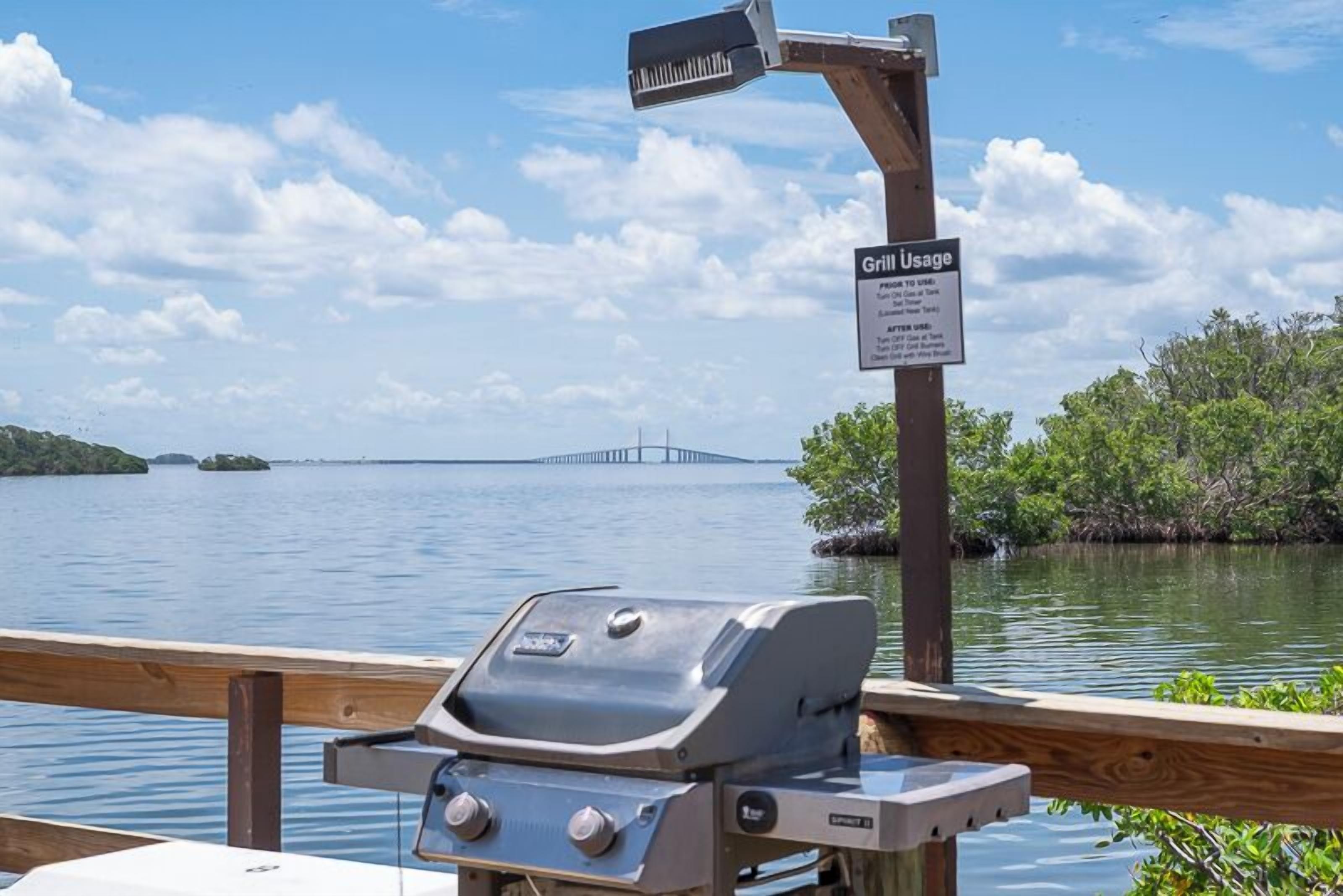 Un barbecue avec une pancarte indiquant son utilisation, une rambarde en bois et une vue sur un pont enjambant une étendue d’eau.