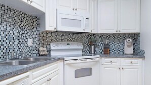A kitchen with white cabinets, a white stove, a microwave, and a tiled backsplash. - Condos at Wander Residences in Tierra Verde (Tierra Verde)