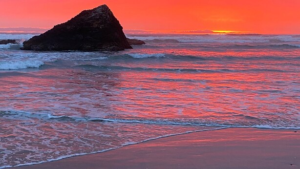 Uma praia com ondas e uma formação rochosa ao pôr do sol.