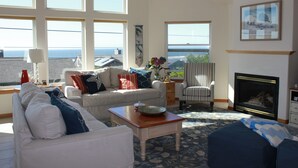 Sala de estar com sofá, poltrona e lareira. Janelas amplas oferecem vista para o mar.