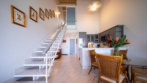 Una cocina moderna con armarios blancos, un comedor con sillas de mimbre y una escalera que sube a un nivel superior.