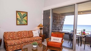 Una sala de estar con sofá, sillón y una mesa con una planta. Se ve una ventana con vista al mar.