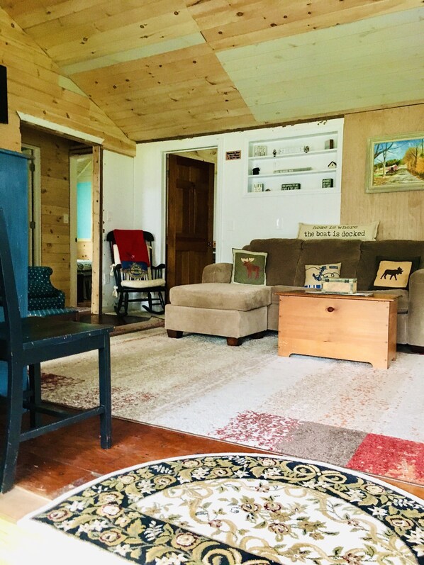 A cozy living room with a wooden ceiling, a sofa, a chair, a small table, and a patterned rug. - Quaint Cottage On The Lake! (Hague)