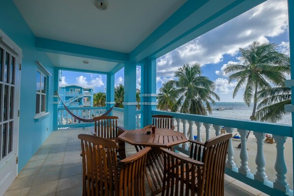 A turquoise room with a wooden table and chairs, a hammock, and a view of the ocean and palm trees. - Sunset Beach Resort "B2" - Beachfront, Free Kayaks, Bikes & More! 🏖 (San Pedro Town)