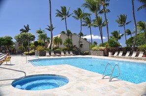 A swimming pool with lounge chairs and a hot tub, surrounded by palm trees and a building.