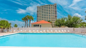 Una piscina con sillas de descanso, un edificio con techo rojo y un hotel alto al fondo.