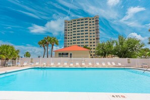 Piscina com espreguiçadeiras, um edifício com telhado vermelho e um hotel alto ao fundo.