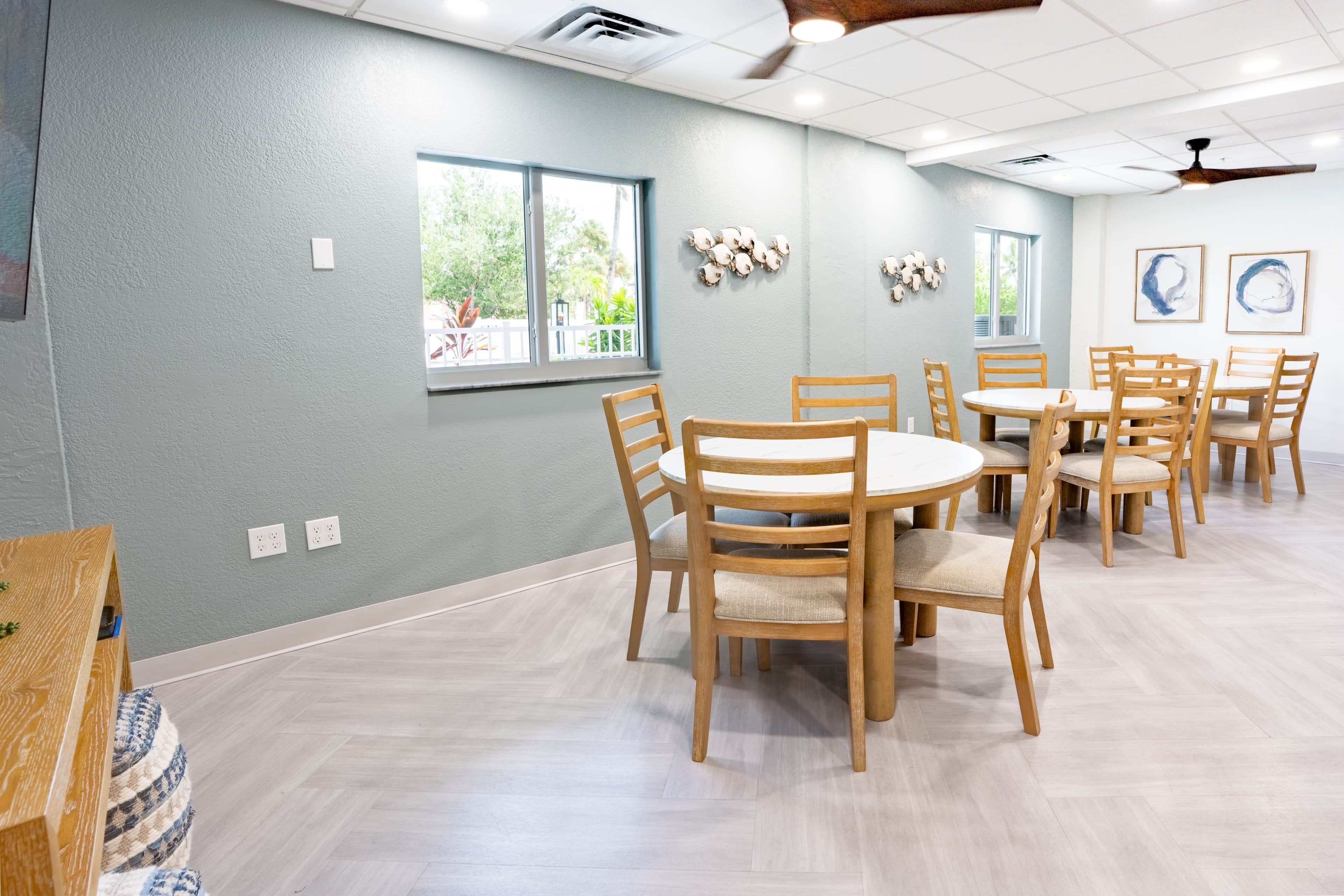 Ein Zimmer mit einer hellgrauen Wand, Holztischen und -stühlen, einem Deckenventilator und Fenstern mit Blick auf Grünflächen.