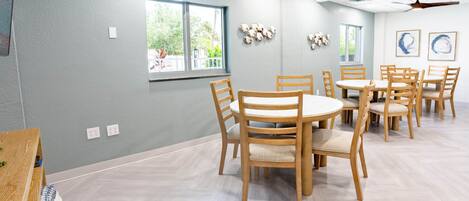 Ein Zimmer mit einer hellgrauen Wand, Holztischen und -stühlen, einem Deckenventilator und Fenstern mit Blick auf Grünflächen.