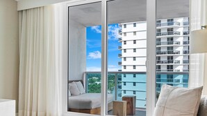 Una sala moderna con un gran ventanal corredera que da a un balcón, un sofá de mimbre y vistas de un edificio de varios pisos.