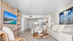 Una sala de estar moderna con una silla de mimbre, un sofá blanco y una mesa de centro. Se observa una cocina al fondo y un cuadro grande en la pared.