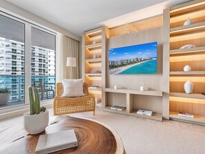 A modern living room with a large window, a wicker chair, a flat-screen TV, and built-in wooden shelves. - Luxurious 1/1 Ocean View Condo 1009 - Located at 1 Hotel & Homes South Beach (Miami Beach)