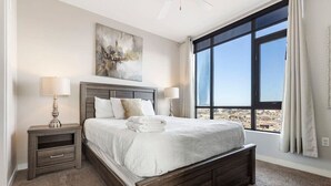 Ein Schlafzimmer mit einem großen Fenster, einem Bett mit weißen Leinen, zwei Nachttischen mit Lampen und einem Gemälde an der Wand.