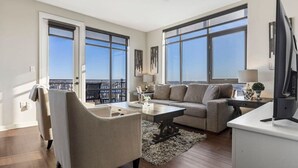 Ein geräumiges Wohnzimmer mit einer Couch, einem Sessel, einem Couchtisch und einem Fernseher. Große Fenster bieten Blick auf die Stadt.