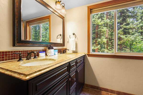 Un baño con lavamanos de encimera de granito, muebles de color oscuro, un espejo grande y una ventana con vistas a un bosque.
