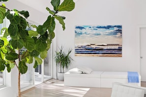 A modern living room with a large window, a white sofa, and a beach painting on the wall. - Contemporary East Hampton Retreat, Tennis + Pool (East Hampton)