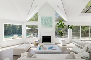 A spacious living room with a fireplace, large windows, and a view of an outdoor pool. - Contemporary East Hampton Retreat, Tennis + Pool (East Hampton)