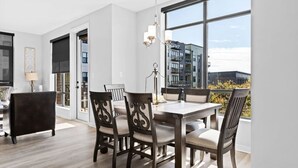 Ein Essbereich mit Tisch, Stühlen, einer Lampe und Blick auf Gebäude durch große Fenster.