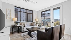 Ein modernes Wohnzimmer mit einer Couch, einem Couchtisch und Sesseln. Große Fenster und ein Deckenventilator sind vorhanden.