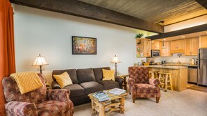 A cozy living room with a sofa, two patterned armchairs, a coffee table, and two lamps. A kitchen with wooden cabinets and a stainless steel refrigerator is visible in the background. - Pool, Hot Tub, Weber Gas BBQ, Renovated. Easy access into unit (Steamboat Springs)