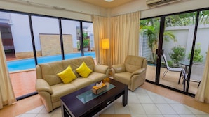 A living room with a beige sofa, a matching armchair, and a glass-top coffee table. There is a sliding glass door leading to a pool area.