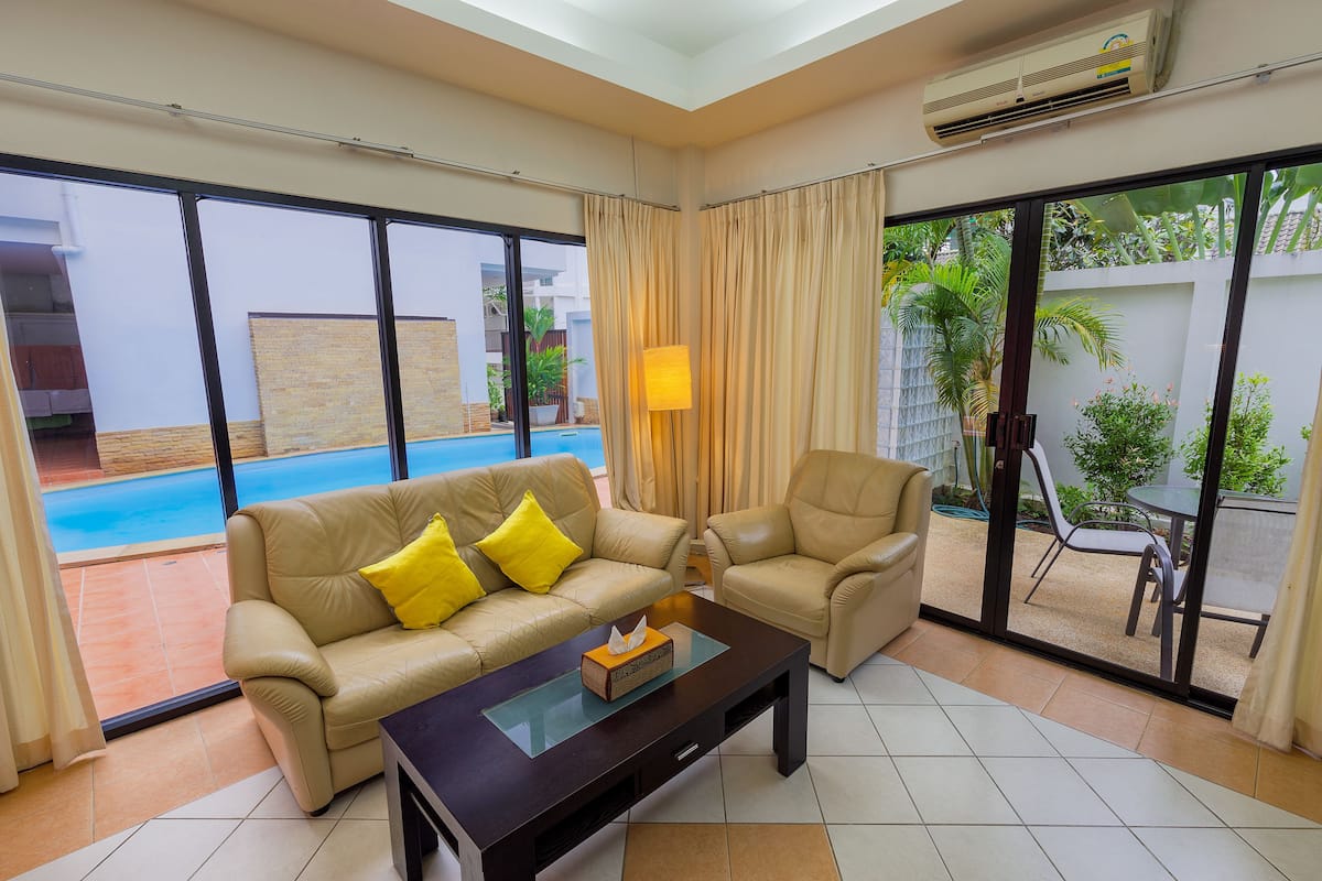 A living room with a beige sofa, a matching armchair, and a glass-top coffee table. There is a sliding glass door leading to a pool area.