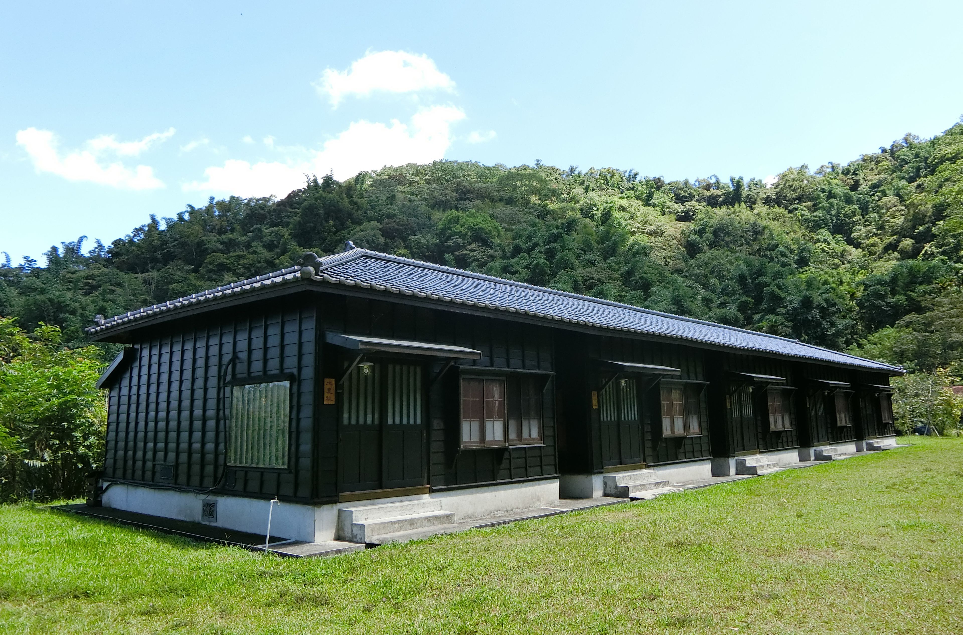 Un edificio japonés tradicional con tejado de tejas, situado en una zona verde exuberante con una colina boscosa al fondo.