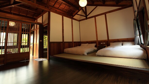 Ein traditioneller japanischer Raum mit Tatami-Böden, einer niedrigen Plattformliege, Holzbalken und einer Shoji-Schiebetür.