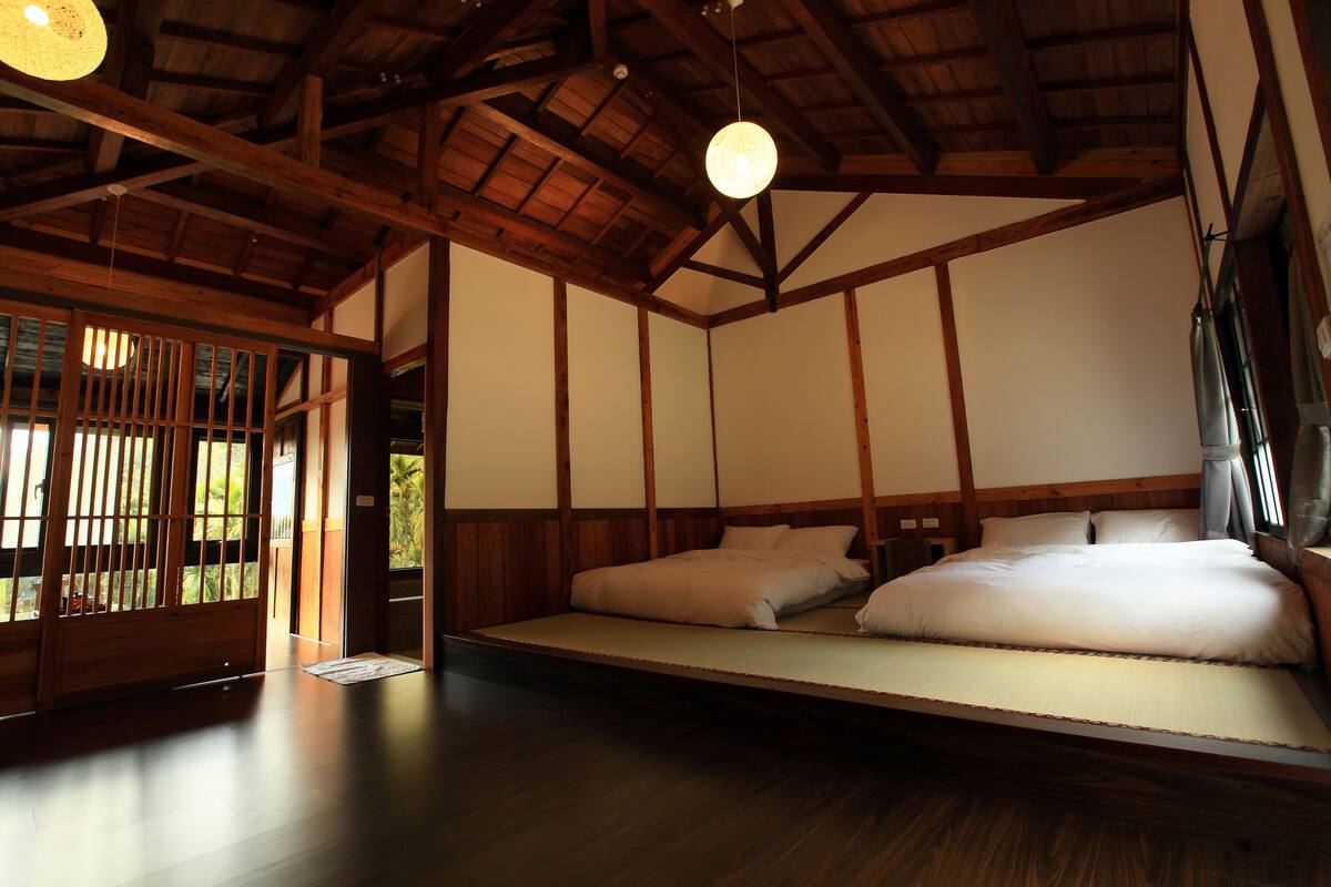 Une pièce de style japonais traditionnel, avec un sol recouvert de tatamis, un lit bas sur une plateforme, des poutres en bois et une porte coulissante de type shoji.