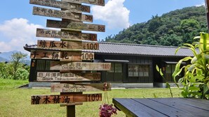 Ein hölzernes Wegweiser mit verschiedenen Zielen und Entfernungen, ein traditionelles Gebäude und eine gebirgige Landschaft.