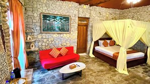 A stone-walled room with a red sofa, a bed with a canopy, a small table with a tray of items, and a stone floor. - RAJAKLANA Resort, Villa And Spa (Bangunjiwo)