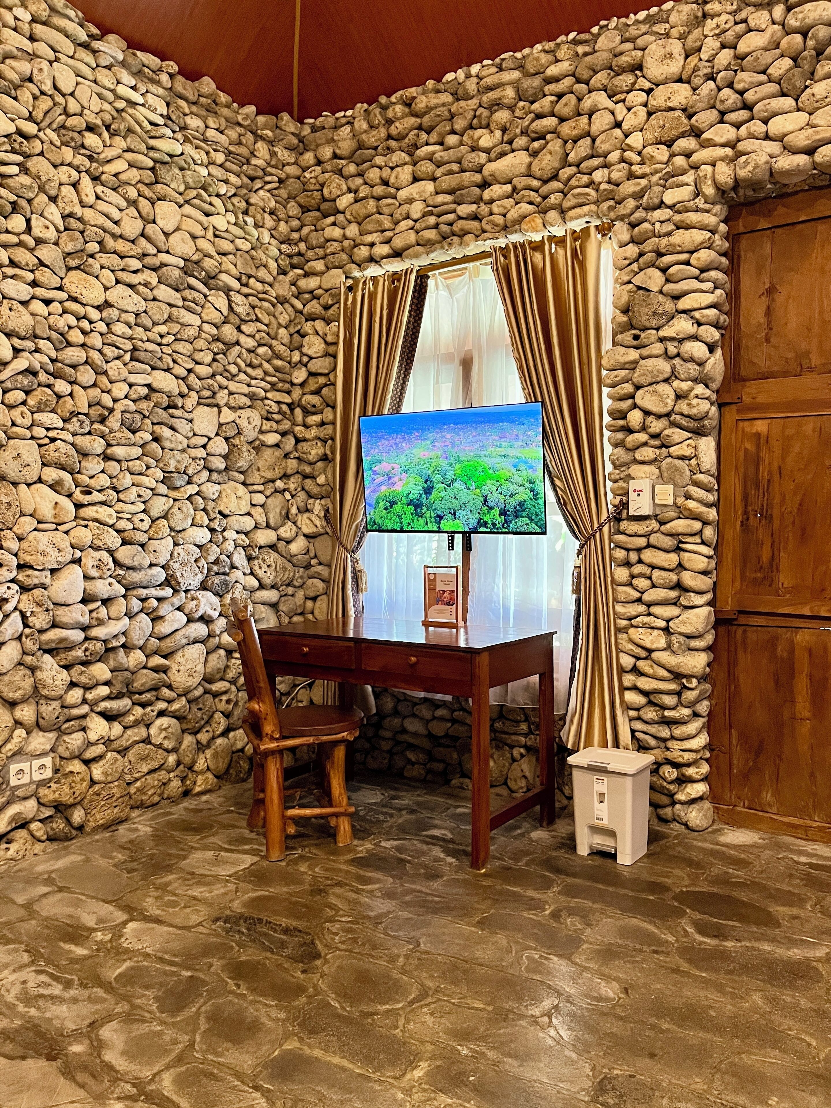 Habitación con paredes de piedra, un escritorio de madera, una silla y un televisor que muestra un paisaje.