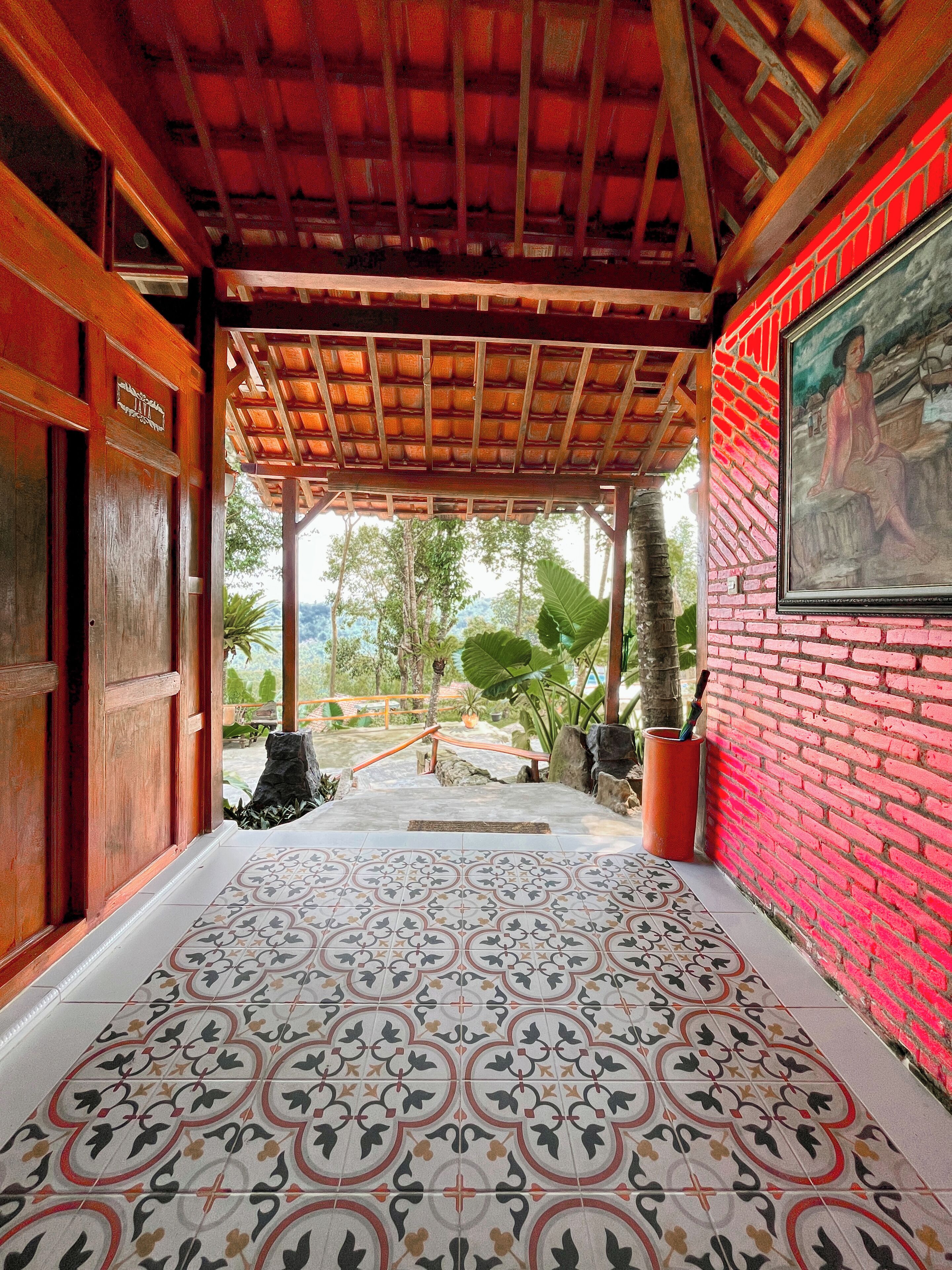 A covered walkway with patterned flooring, a wooden door, a brick wall with a framed picture, and a view of greenery.