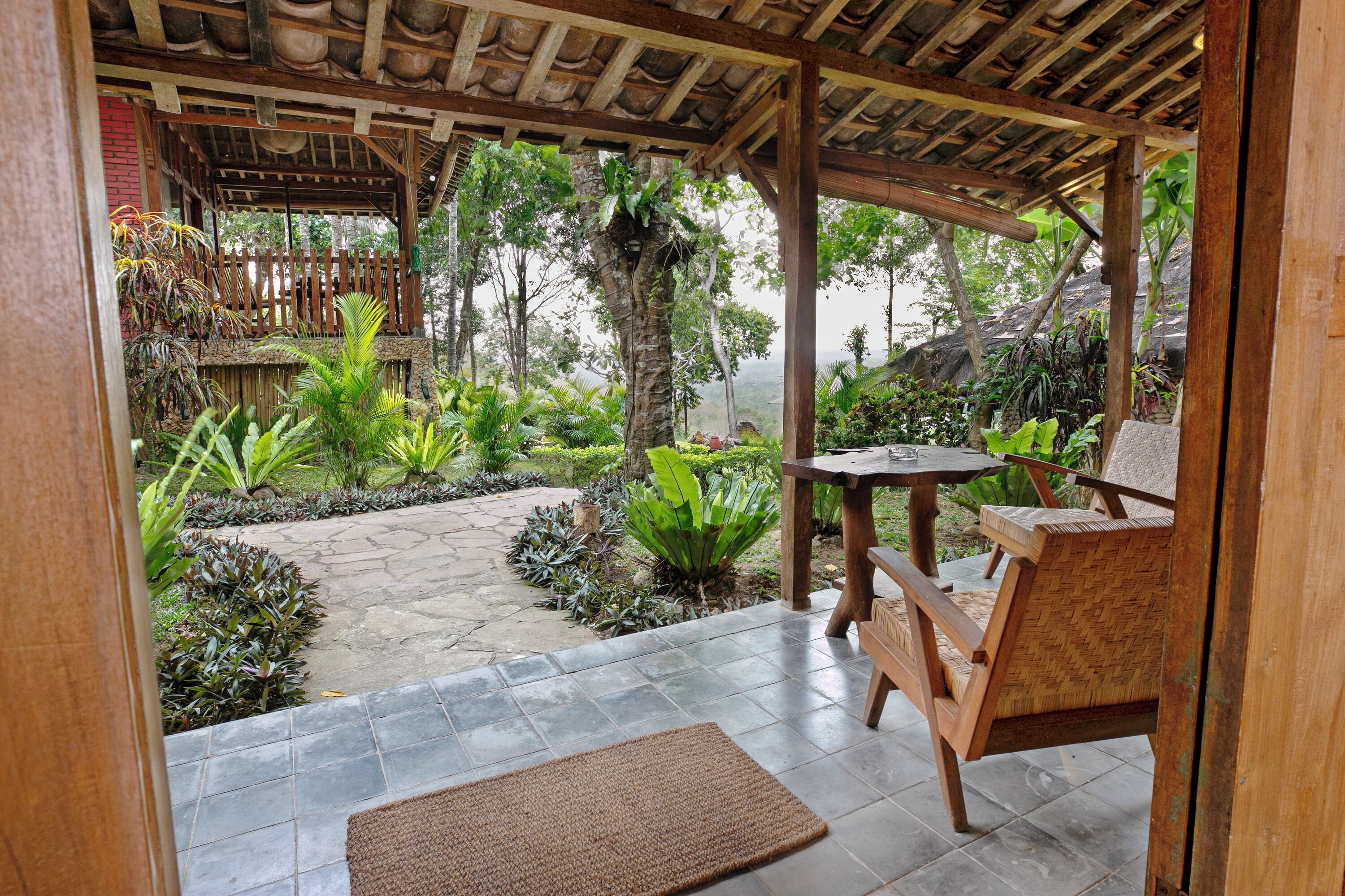 A covered outdoor area with a wooden bench and table, surrounded by lush greenery and a stone pathway.