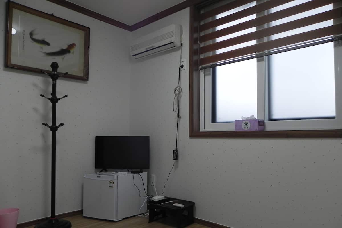 A room with a flat-screen TV, a small refrigerator, a pink trash bin, and a window with blinds.