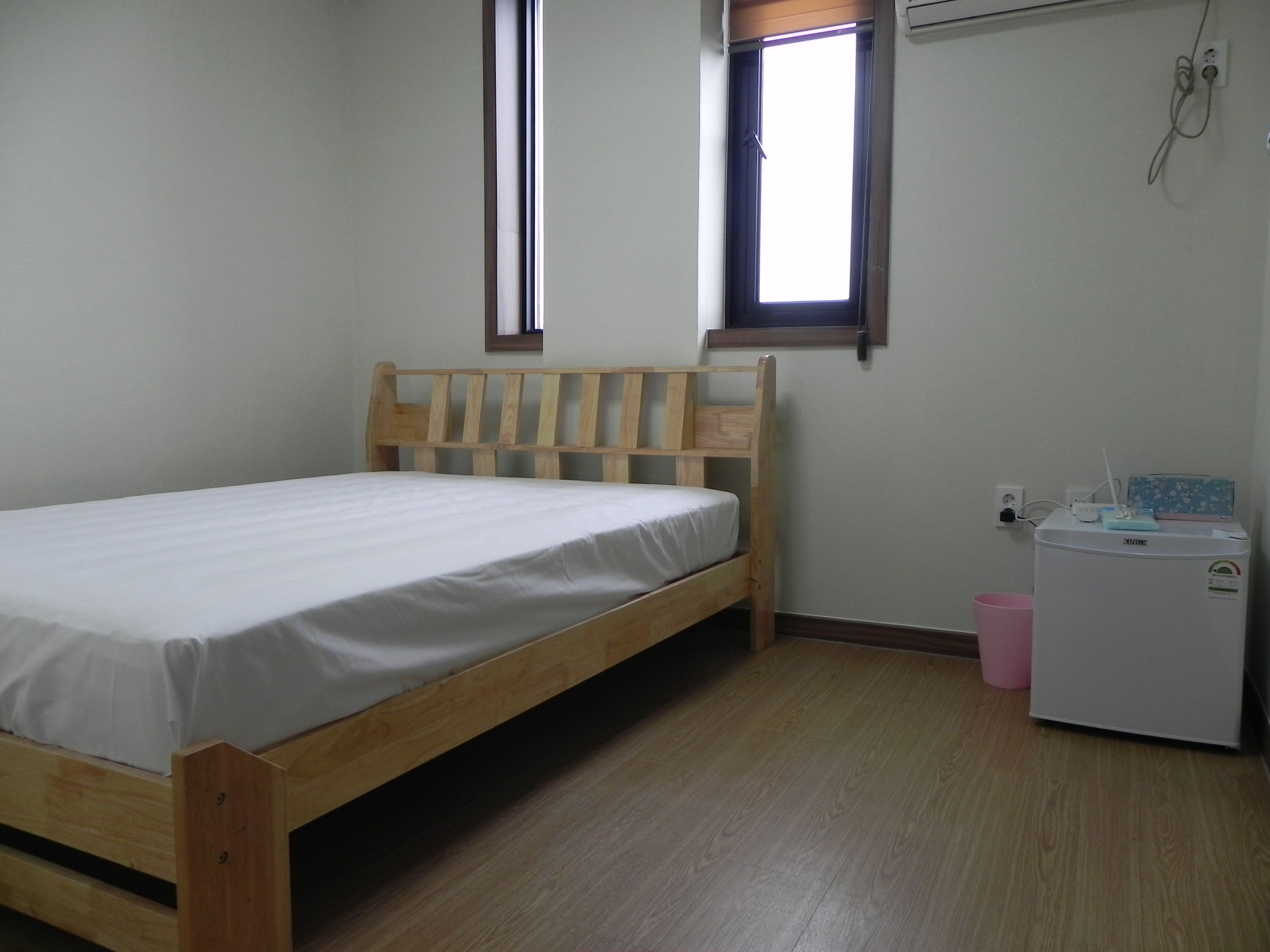 A single bed with a wooden headboard in a room with a small window, a wall-mounted air conditioner, and a compact refrigerator.
