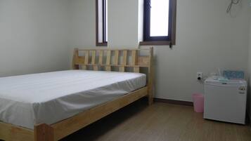 A single bed with a wooden headboard in a room with a small window, a wall-mounted air conditioner, and a compact refrigerator.