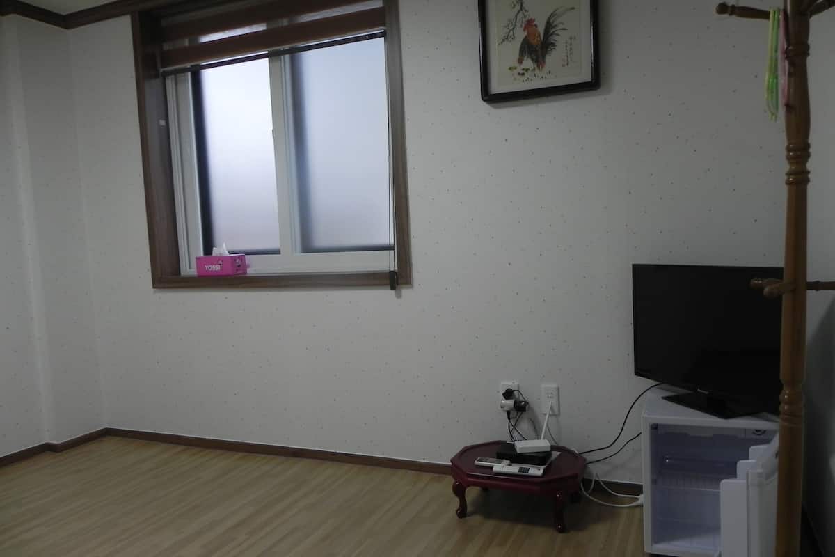 A room with a large window, a television, a small table with a remote control, and a refrigerator.