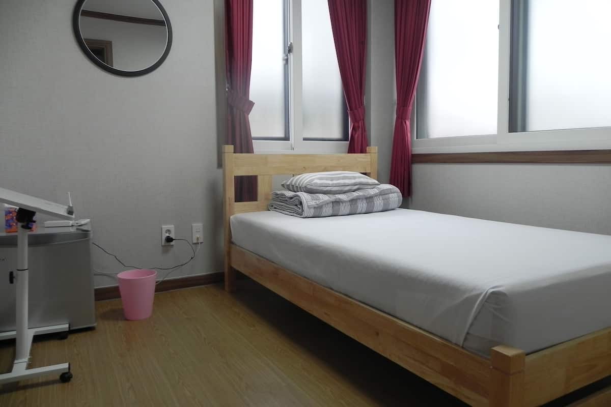 A single bed with a wooden frame and white bedding, a round mirror on the wall, and a window with red curtains.