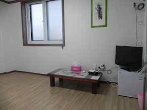 Una habitación con piso de madera, una mesa con una caja de pañuelos rosas, un televisor negro sobre un soporte y una ventana con persianas.