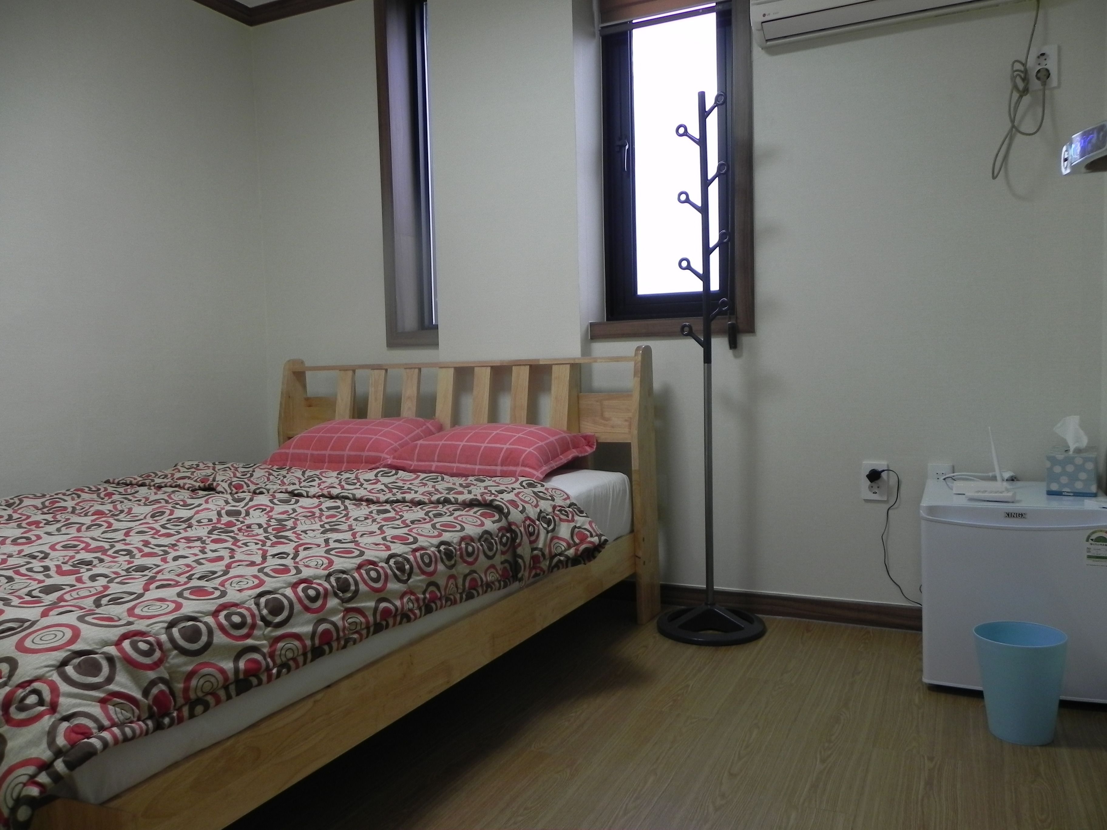 Ein Schlafzimmer mit einem hölzernen Bett, gemusterten Betttextilien, einem Nachttisch, einem kleinen Schreibtisch, einer blauen Mülltonne und einem Fenster mit einem Metallgestell.
