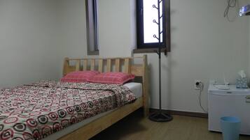 Ein Schlafzimmer mit einem hölzernen Bett, gemusterten Betttextilien, einem Nachttisch, einem kleinen Schreibtisch, einer blauen Mülltonne und einem Fenster mit einem Metallgestell.