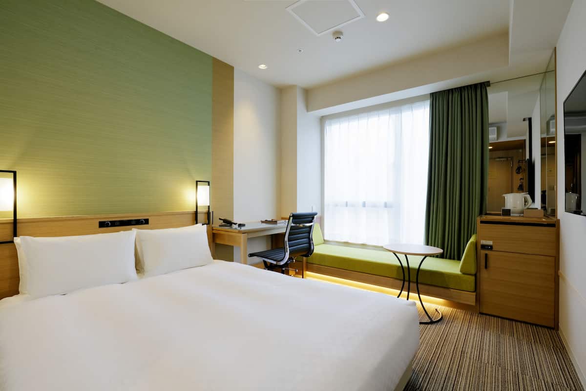 A hotel room with a large bed, a desk with a chair, a small table, a green sofa, a wooden cabinet, and a window with curtains.