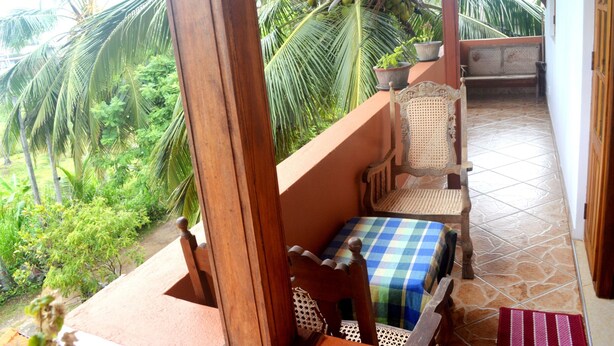 A balcony with a wooden chair and a small table, overlooking a lush garden with palm trees. - Shyni Tourist Guesthouse (Hikkaduwa)